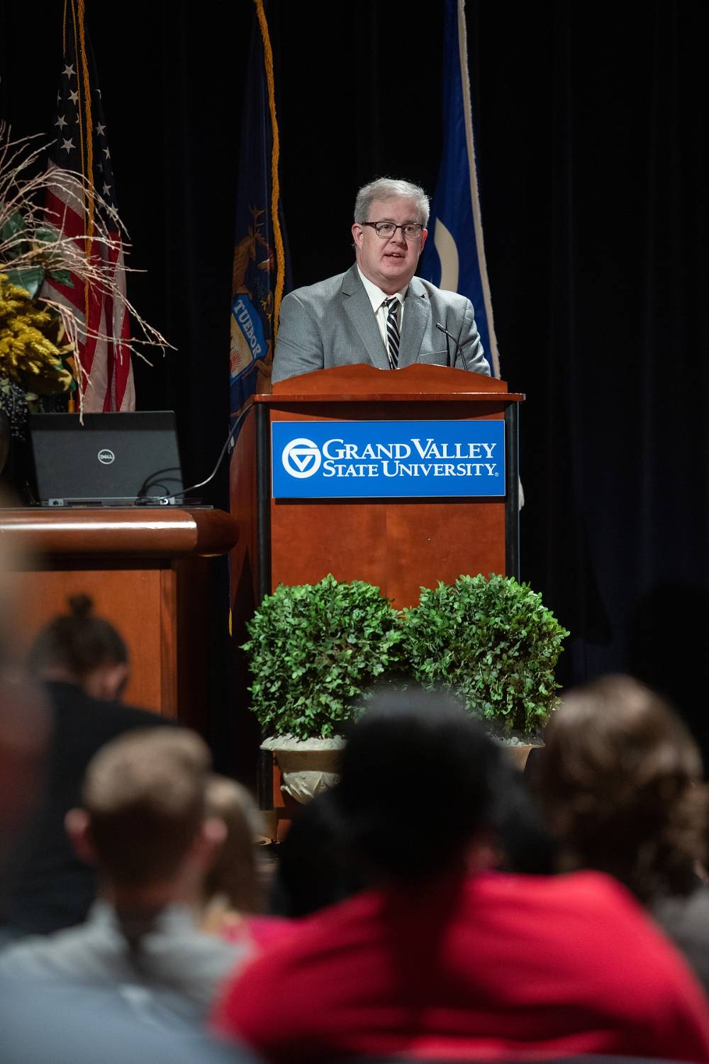 Man stands talking at a GVSU podium, with people sitting out of focus in the foreground